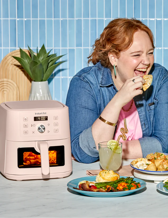 A woman with red hair, wearing a denim shirt and pink top, laughs while eating a snack at a table set with plates of food and a drink. Next to her is the Instant Pot® 4QT VORTEX® Plus Mini Air Fryer in Rosewater.